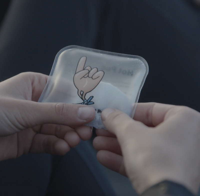 A close-up of a person's hands holding a translucent square patch or sticker with a cartoon hand graphic and partial text, against a dark blurred background.