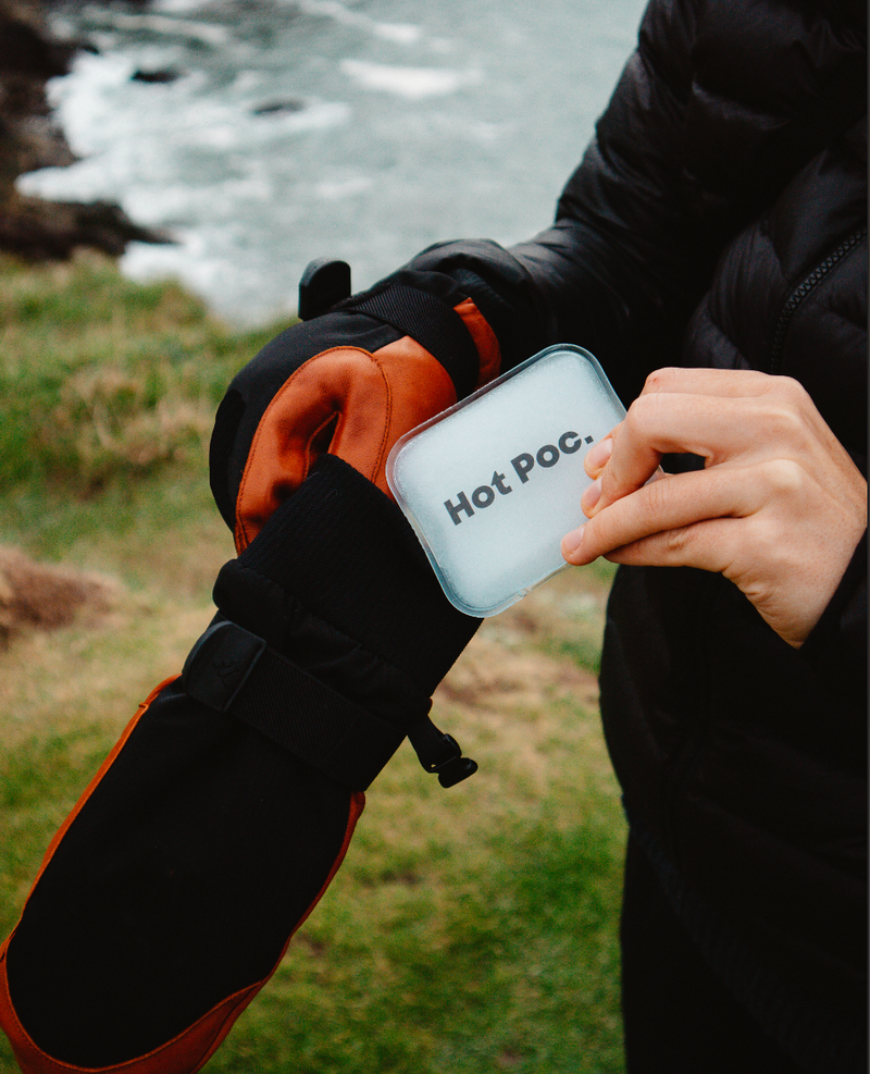 A person outdoors by the water wearing a black and orange winter glove while holding a small square Hot Poc. insulated cup against the glove.
